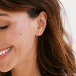 A close-up of a woman with brown hair wearing a small, delicate silver stud earring on her earlobe.