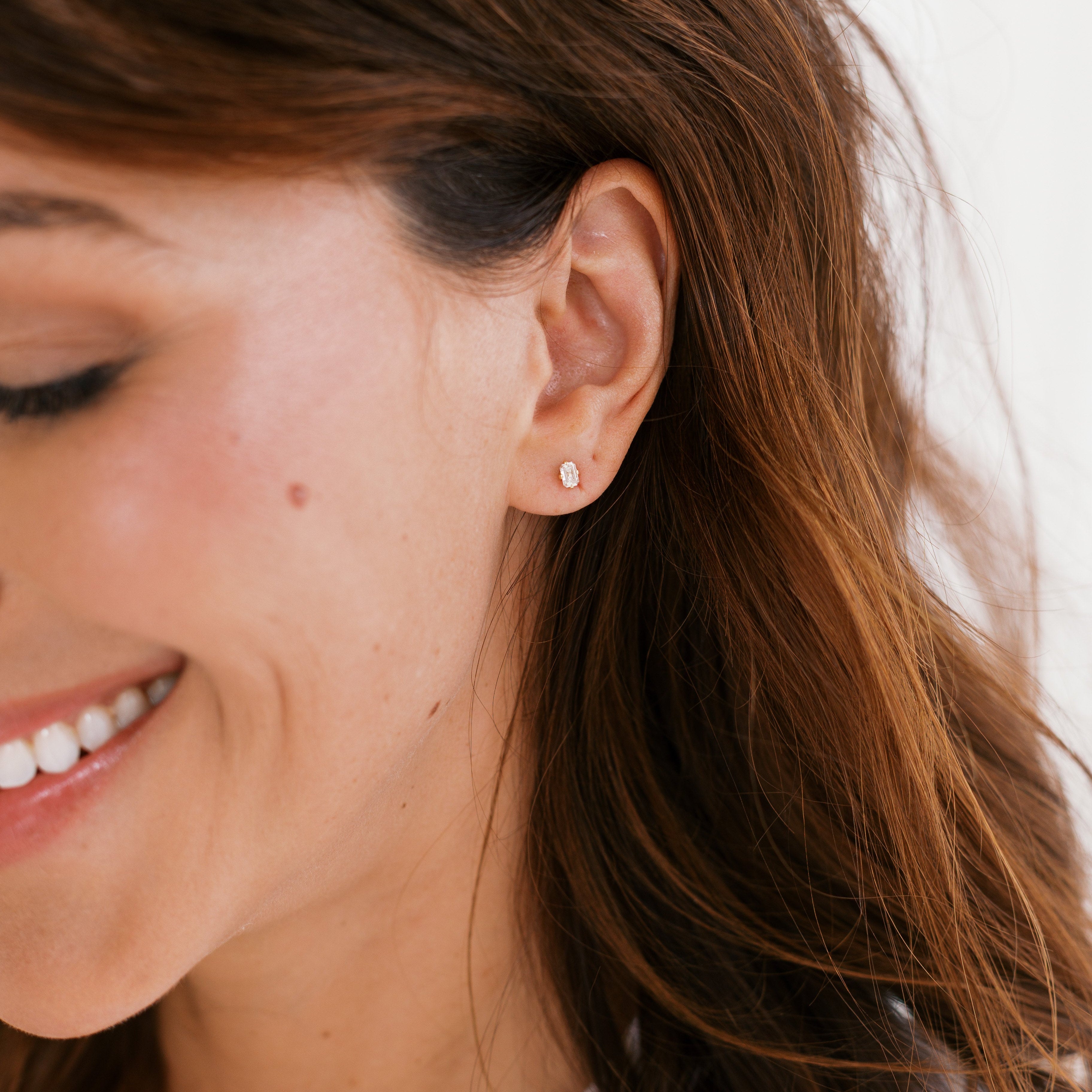 A close-up of a woman with brown hair wearing a small, delicate silver stud earring on her earlobe.